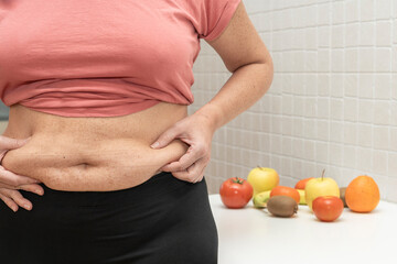 Middle-aged woman shows excess abdominal fat and in the background various fruits and vegetables....