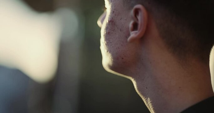 A young man with acne on his face is looking down backlit by the sunset setting sun. Concept of sadness and loneliness, cinematic isolated shot