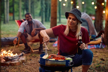 Young woman preparing food on barbecue grill while camping in woods.
