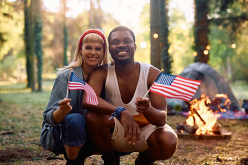 Happy couple with flag of United States of America camping in woods and looking at camera.