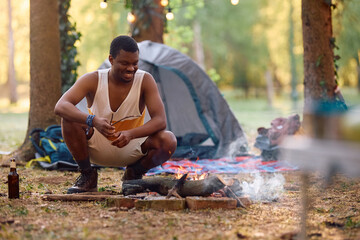 Happy black man setting campfire in woods.