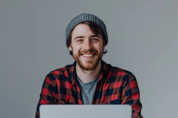 A young cheerful man in a beanie is smiling while working or surfing on his laptop computer