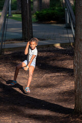 Young girl smiling on swing, dappled sunlight, park setting, wearing blue shorts and sandals, touching her hair.