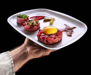 Chef holds white plate of steak tartare on black background. Tasty food background.