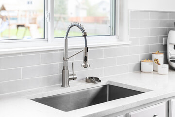 A kitchen faucet detail with a stainless faucet and sink, grey subway tile backsplash, and...