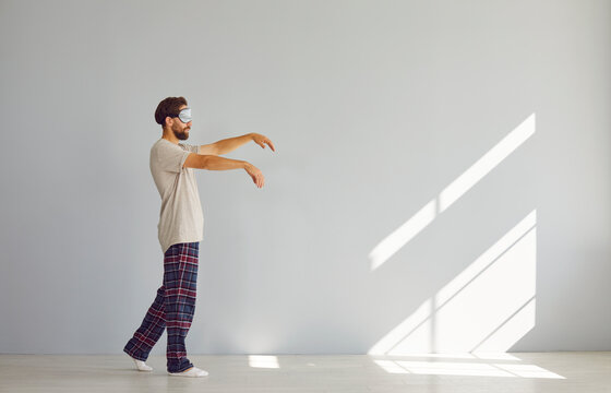 Full length portrait of young sleepwalker man in pajamas suffering from sleepwalking in sleep mask isolated on gray wall background. Somnambulism, insomnia and sleep problems concept.