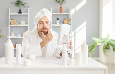 Man is taking care of facial skin, using a mirror and face creams while sitting at a table after a...