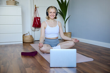 Portrait of fit and healthy woman at home, practice yoga, sitting on rubber mat, listening to...