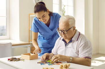 Friendly caregiver or nurse in the retirement home helping a senior man with puzzle games. Demented old man sitting at a desk and solving wooden alphabet puzzle. Dementia, Alzheimer's disease concept