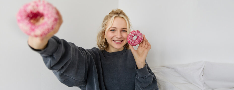 Portrait Of Beautiful Blond Woman, Showing Two Pink Doughnuts, Delicious Pastry, Eating Dessert And Smiling, Sharing Food With You