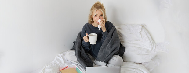Portrait of young woman sitting at home sick, feeling unwell, drinking tea, spending time in bed,...
