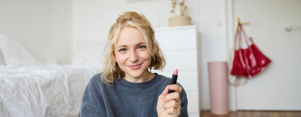 Portrait of smiling beautiful woman in her room, sitting and showing lipstick, recommending...