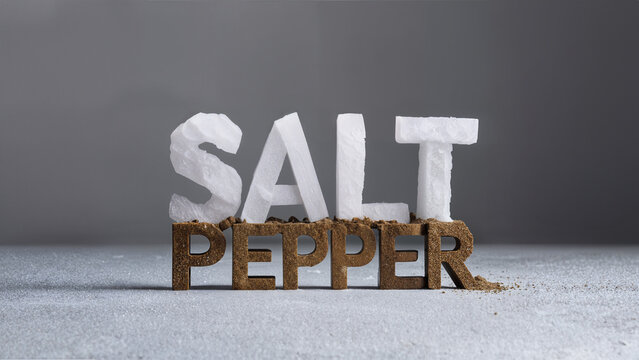 Large salt crystal and pepper powder letters forming the word 'salt pepper' on a minimal grey backdrop and sprinkled salt surface.
