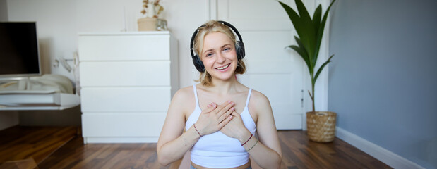 Portrait of young happy woman in headphones, looking with relaxed, friendly face expression, holding hands on her chest and smiling