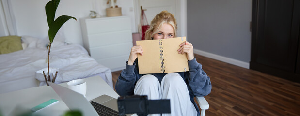 Portrait of cute blond woman, sits in front of digital camera and laptop in her room, covers face...
