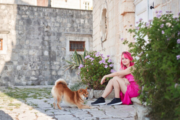 A woman with vibrant pink hair gently pets a Shiba Inu dog in a quaint stone-paved alley, a tender moment of connection.
