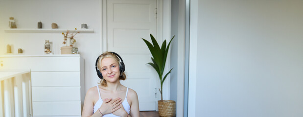 Vertical portrait of beautiful young woman in headphones, sits on yoga mat, meditating, holding hands on her chest and smiling at camera