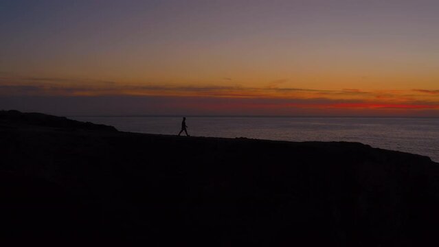 Silhouette of man walk on edge of cliff at epic sunset. Breathtaking view of the burning horizon over the ocean. Incredible inspiring background footage with copy space.