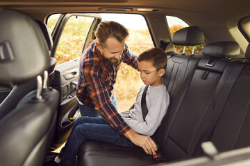 Caring father is buckling up seat belt for his young son in back seat of family car. Man helps...
