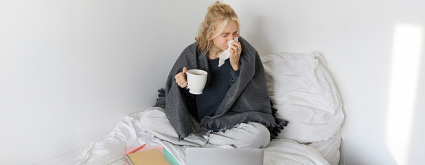 Portrait of woman catching a flu, sneezing, feeling sick, sitting on bed with laptop and working on...