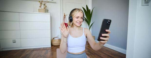 Portrait of young woman with smartphone and headphones, waving hand at mobile phone camera, live streaming during workout