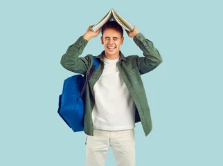 Foto op Plexiglas Happy college or high school student having fun with book. Studio portrait of funny cheerful boy with backpack standing isolated on light background holding textbook on top of his head and laughing © Studio Romantic