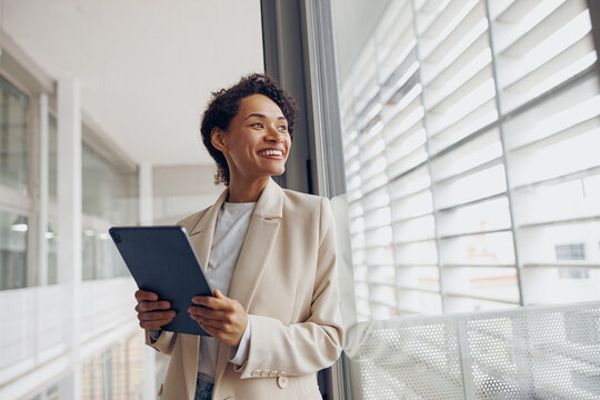 Pretty female manager with digital tablet standing on modern office background and looks away - Powered by Adobe