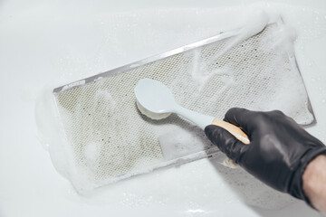 Hand in black glove cleaning mesh filter of cooker hood with brush and foam, white background