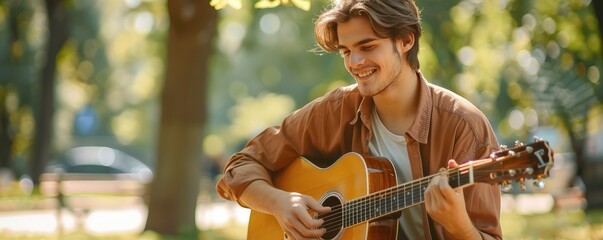A male musician plays acoustic guitar in a park bathed in sunlight amidst green trees.