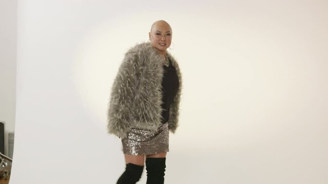 Woman with a bald head dances on a white background wearing fur and sparkles