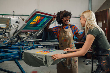Smiling interracial print shop female workers silkscreen printing t-shirt.