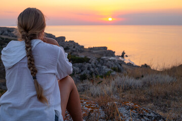 A traveler woman sits on the mountain and looks at the sunset over the sea.