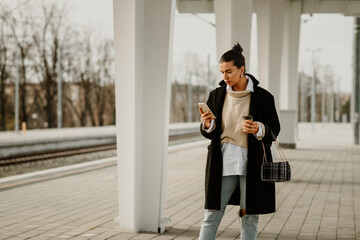 A fashionable woman standing on train station and typing on her phone.