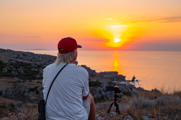 A traveler man sits with a video camera on the mountain and removes the sunset over the sea.