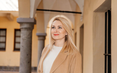 Fototapeta premium Elegant Woman in a Beige Coat Smiling Softly in an Arched Hallway