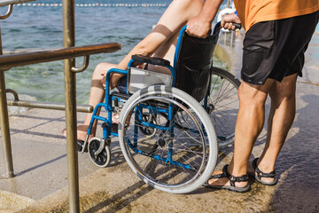Male assistant helping a man in a wheelchair to enjoy the sea on an access ramp into the water. Accessible beach concept.