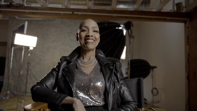 A woman with alopecia smiles at the camera while sitting on a film set