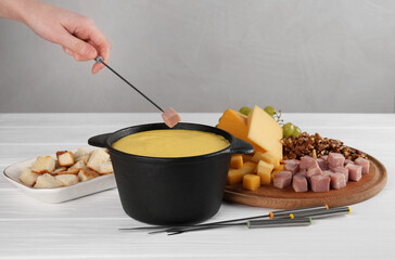 Woman dipping piece of ham into fondue pot with tasty melted cheese at white wooden table, closeup