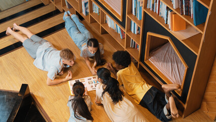Fototapeta na wymiar Group of diverse children lying down in circle while reading a book at library. Top view of girl sharing a magazine while pointing at interested topic and talking with lovely friends. Edification.