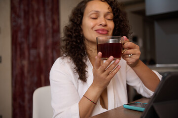 Happy curly haired multi ethnic woman in white waffle bathrobe, smiles relaxing over up of hot tea in home interior. Focus on a glass cup of hot healing herbal drink in women's hands