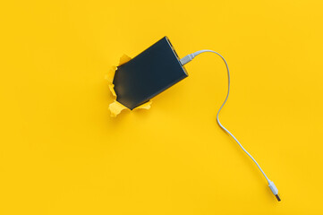 A dark grey metal power bank protrudes from a torn hole in yellow paper. Concept of smartphone...