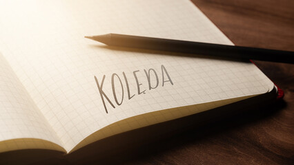 A handwritten inscription "Kolęda" on a grille of an open notebook on a wooden countertop, next to a black pencil, lighting of light. (selective focus), translation: carol
