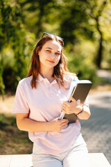 Young cheerful female student is sitting with her books in park or campus on a sunny day.