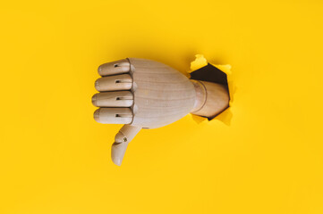 A wooden hand protrudes from a torn hole in yellow paper and shows a thumbs down (dislike). The...