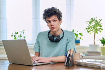 Young male college student sitting at desk with laptop looking at camera - obrazy, fototapety, plakaty