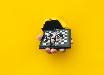 A man's hand comes out of a torn hole in yellow paper and holds a small chessboard with pieces scattered on it. The concept of intellectual development and games that develop thinking.
