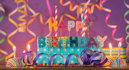 Letters of different colors forming the phrase HAPPY BIRTHDAY, with lit candles against purple...