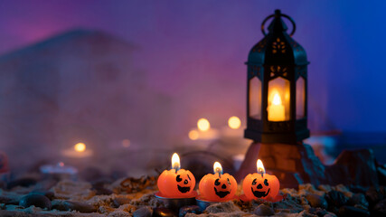 Halloween background, 3 pumpkin-shaped candles in the foreground, an old lamp on a rock against a...