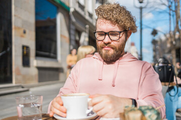 Millennial bearded man having breakfast at table of street cafe on spring day, drinking warm...