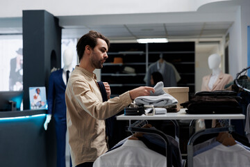 Young man shopping for trendy menswear, browsing through shelf in clothing store. Fashion boutique...